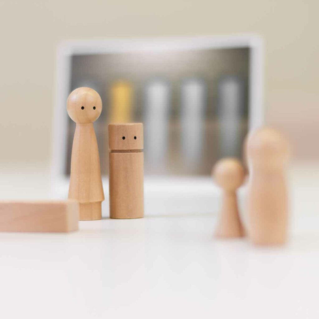 Foto einer mit Holzfiguren aufgestellten Szene: Die große Holzfigur steht im Mittelgrund an der Seite einer kleineren Holzfigur und schaut auf die beiden Figuren im Vordergrund. Den unscharfen Hintergrund bildet eine Fotokarte auf der vier graue und eine gelbe Tür zu sehen sind.