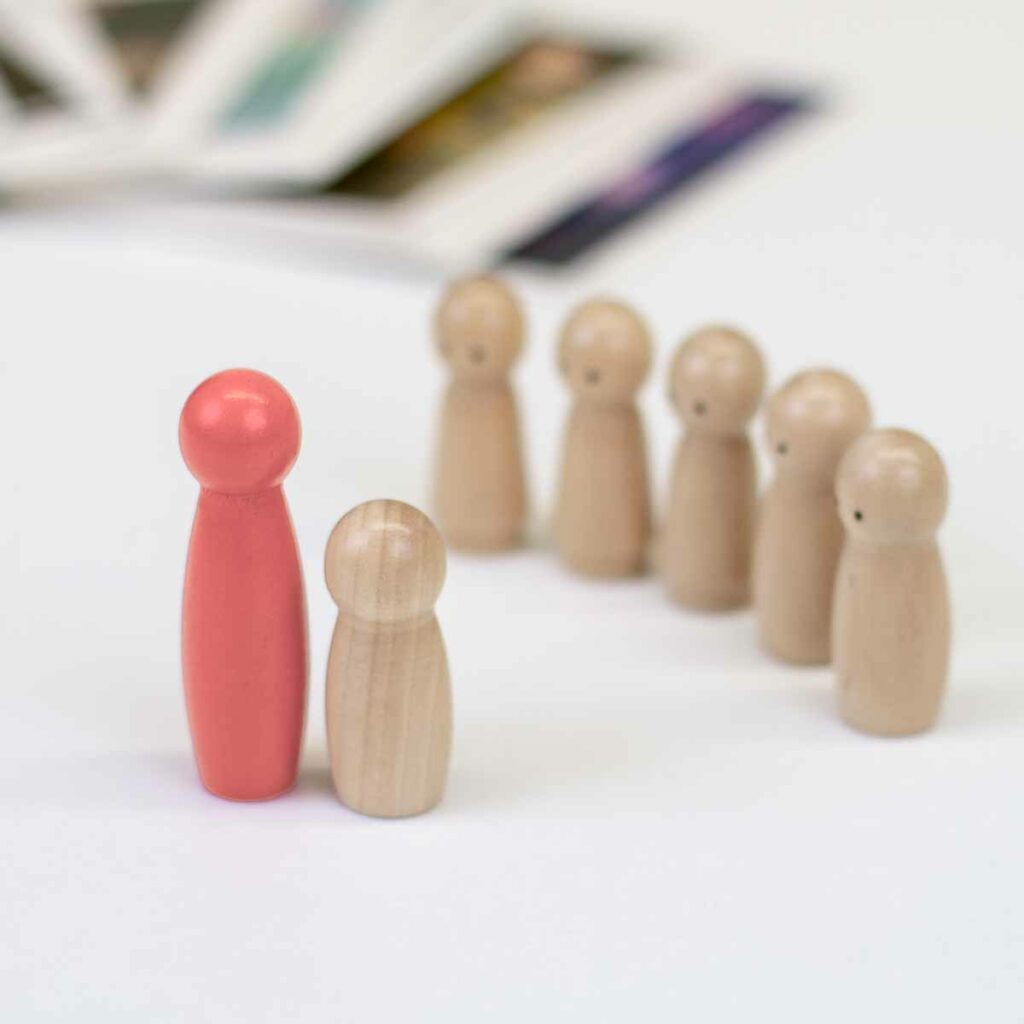 Das Foto zeigt eine mit Holzfiguren gestellte Szene auf weißem Untergrund: Eine große rote Figur steht im Vordergrund neben einer kleineren holzfarbenen Figur. Gegenüber im Halbkreis steht eine Gruppe weiterer Holzfiguren, die alle zur roten Figur schauen.