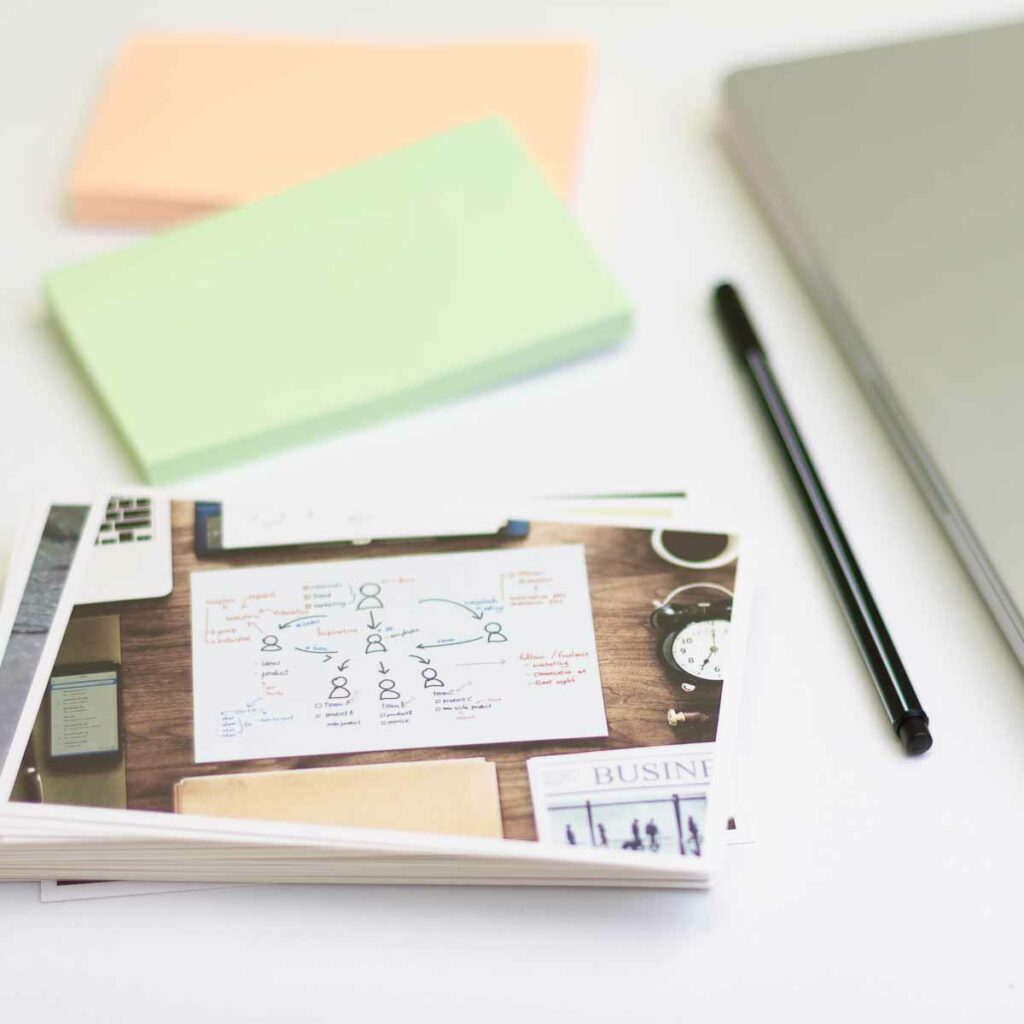 Ein Kartenstapel, ein Stift, zwei Stapel Haftnotizen und ein Macbook auf weißem Untergrund. Die oberste Bildkarte zeigt ebenfall ein Schreibtischmotiv mit einer Bildkarte im Zentrum, auf die mehrere Personen skizziert sind, die (über Pfeile visualisiert) miteinander in Verbindung stehen.